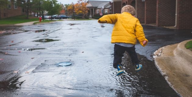 Child in yellow raincoat jumping into a puddle of rainwater on the street.
