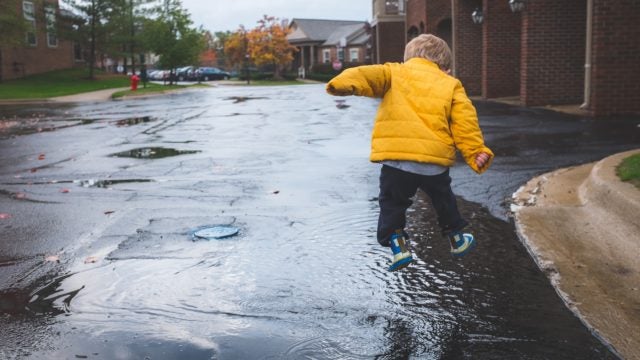 Child in yellow raincoat jumping into a puddle of rainwater on the street.