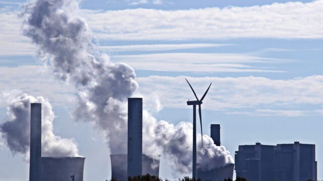 A coal fired power plant spews pollution into the air, while a wind turbine spins in the foreground