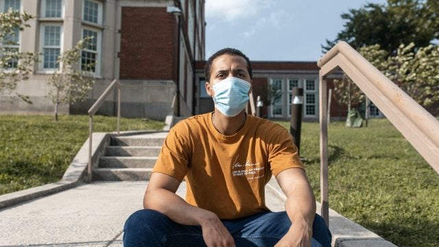 A young Black man wearing a face mask sits on steps outside a brick building