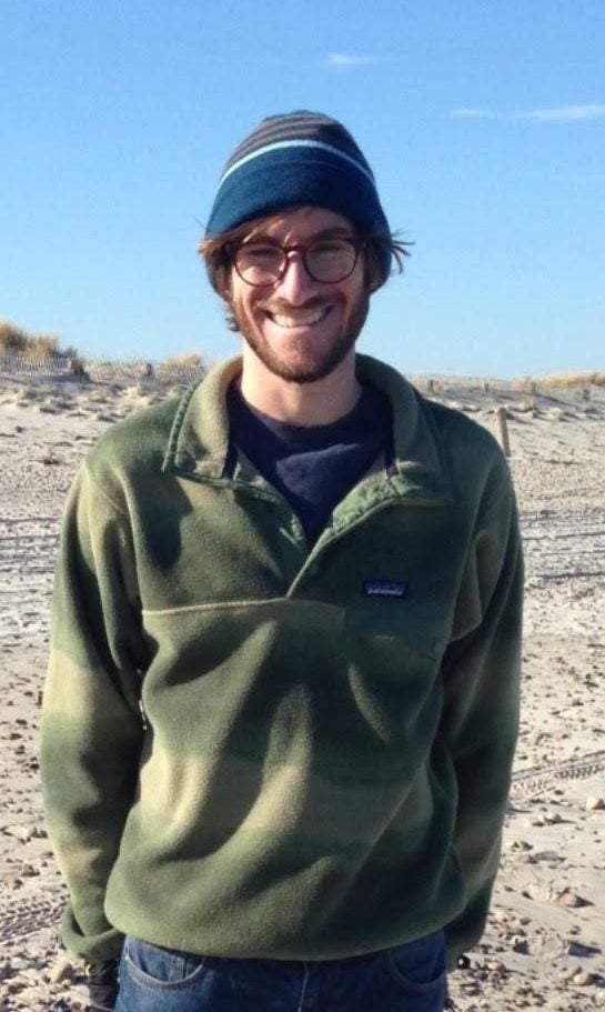 Headshot of Andrew Shapero, wearing a blue beanie and green sweater standing outside.