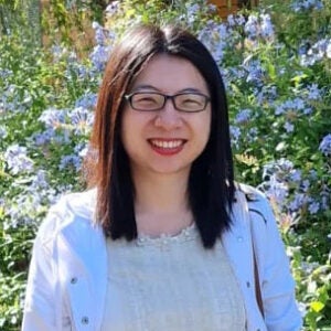 Jiahong in a white jacket, outside in front of blue flowers.