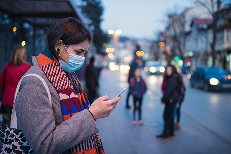 Young woman wearing a face mask looks at her phone at a bus stop.