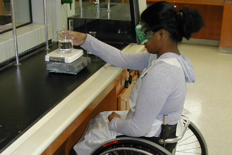 Young woman in wheelchair wearing safety goggles and working at a lab.