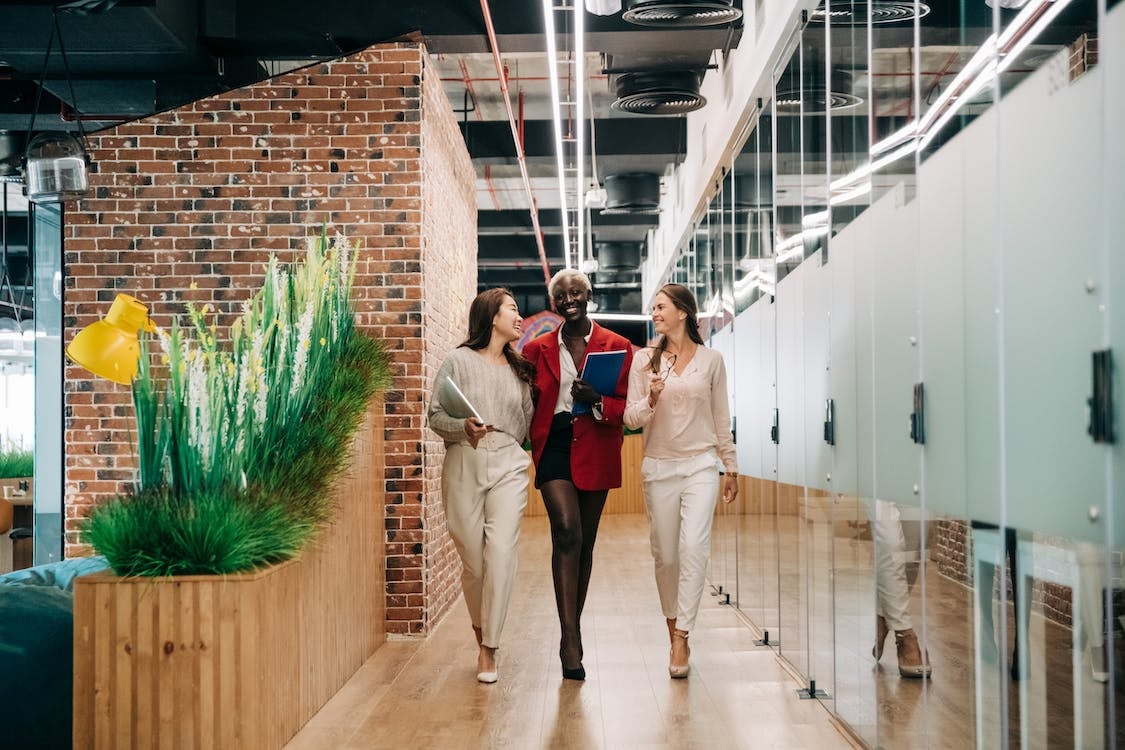 Three business women walking down a hallway in an office building, talking together and smiling
