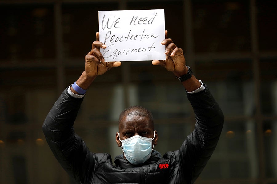 A Black man wearing a mask holds up a sign reading "We need protective equipment"