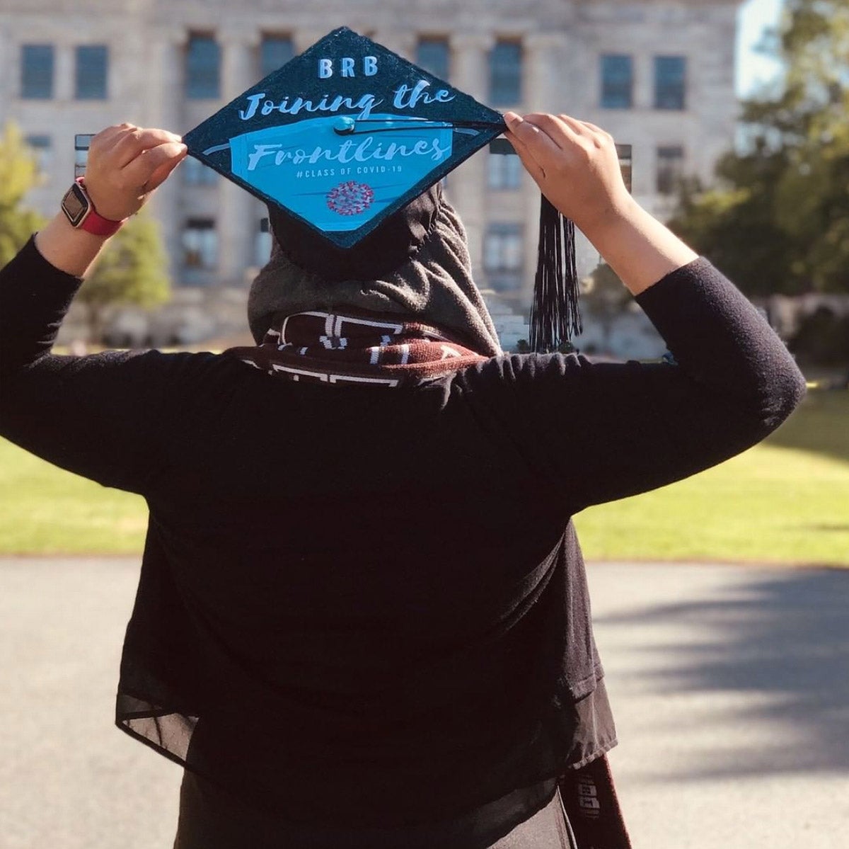 Photo of Taha Khan from behind to show her graduation cap, which reads "BRB Joining the Frontlines"