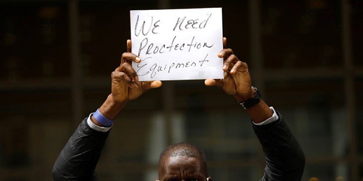 A Black man wearing a mask holds up a sign reading "We need protective equipment"