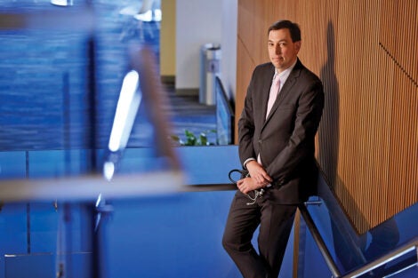 Jesse Ehrenfeld rests on a stairwell railing against a tan wood-paneled wall in a hospital. Blue lights and blue carpet shine on the left side of the image. Ehrenfeld wears a dark suit, light-collared shirt and tie, and holds a stethoscope in his hands.