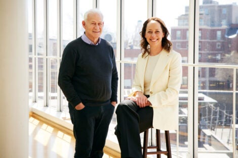 Jack Shonkoff, standing, and Lindsey Burghardt, seated in a stool, pose for a portrait in front of large glass windows in a bright office setting. An urban scene is behind them.