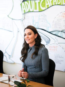 Princess Kate actively listens while sitting at a conference table at the Center on Early Childhood Development. She wears her hair down and wears a black and white checked, collared dress.