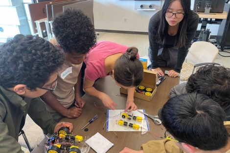 Students and teaching assistants huddle together over a table of toy car parts. One student is attaching a wire to a wheel motor.