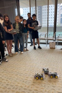 A group of students stands together, some holding open laptops. They are looking at the floor, where three self-driving cars face each other.