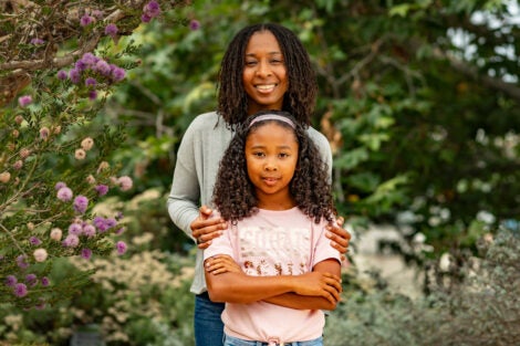 A woman stands behind her young daughter and rests her hands on her shoulders. They book smile at the camera. They are pictured in a lush green park with flowering purple trees in the background.