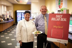 Executive chef Arlene Richburg and Sebastian's manager Paul Doherty stand with a plate of food in the cafeteria next to the Daily Dollar Deals sign