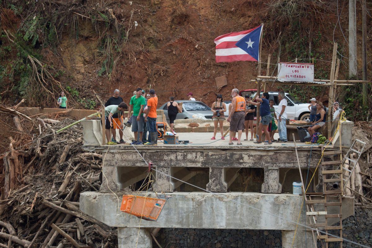 Utuado, Puerto Rico -- Villagers collect on bridge remnants to receive needed supplies. The village has been cut-off by the Rio Charco Abajo's flooding that took away the village road and bridge. An ingenious cable pulley system ferries a shopping cart from shore-to-shore.