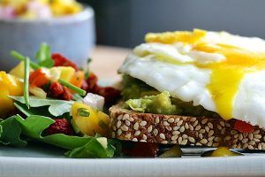 Poached egg with runny yolk on mashed avocado and whole grain bread, with a side salad of arugula and mango red pepper salsa