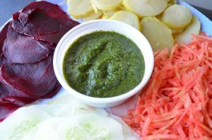 a bowl of green chutney surrounded by sliced vegetables including beets potatoes carrots lettuce