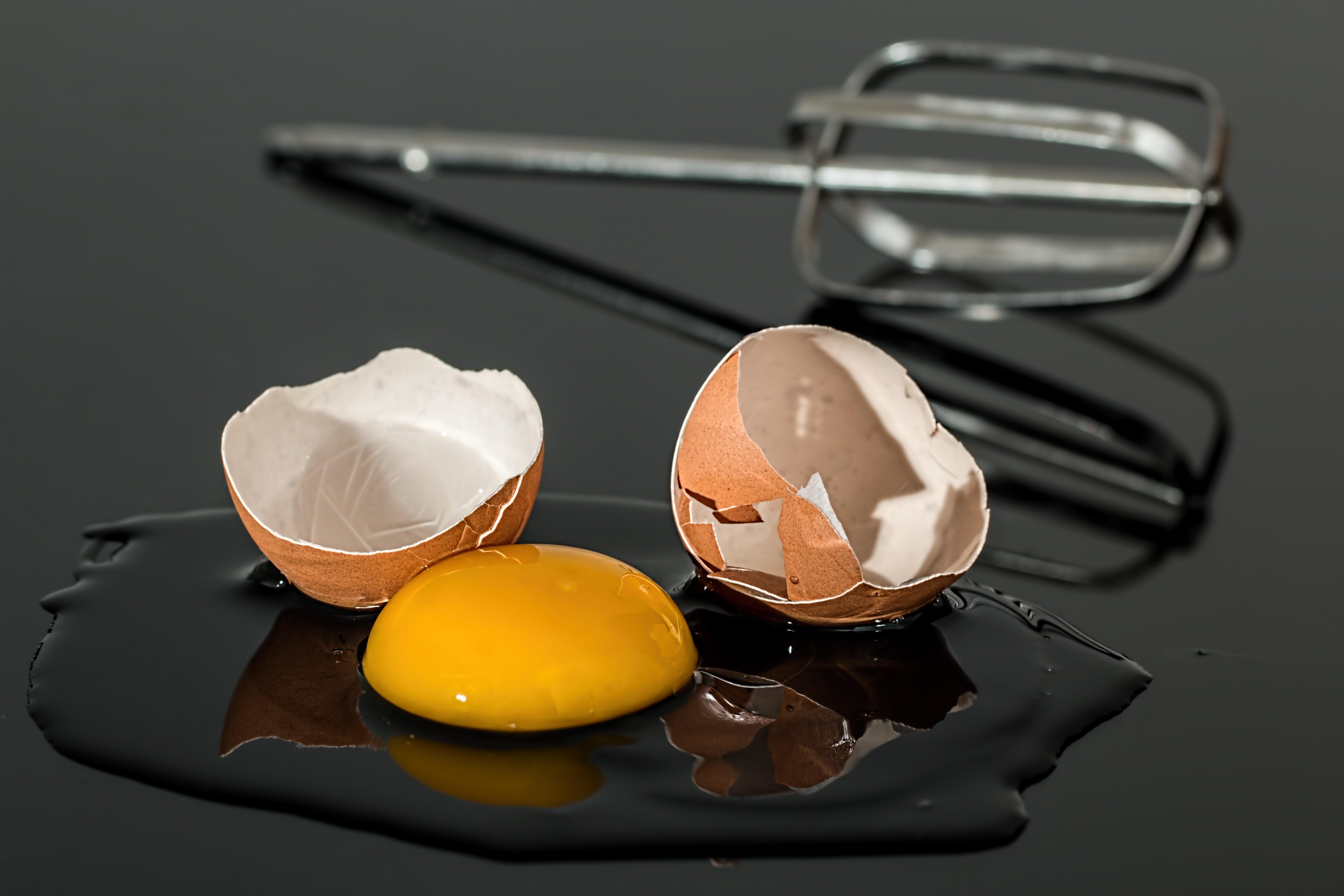 Cracked egg on a table with yolk and eggshells, whisk