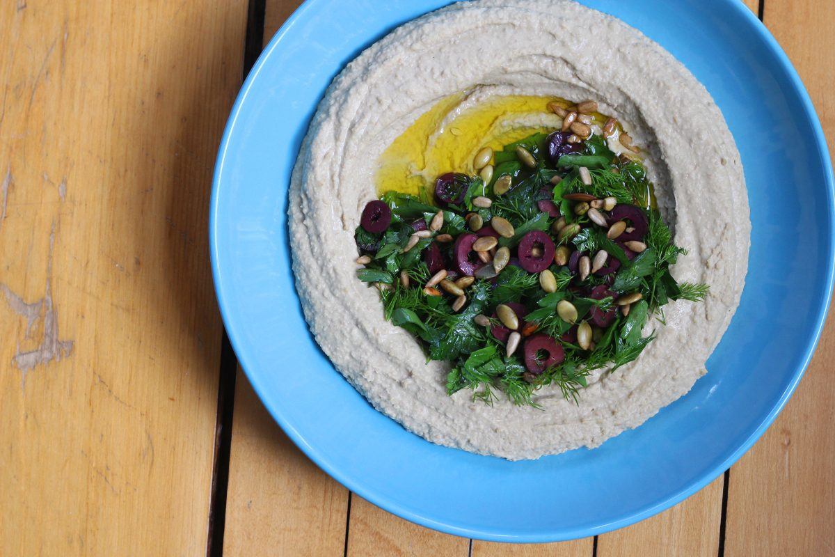 green lentil hummus with a variety of herbs, seeds, and olives, on top of a blue plate on a table