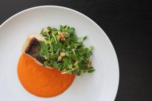 A plate with pan seared bass fish over a bright red walnut romesco sauce and topped with a bright green pea shoot salad