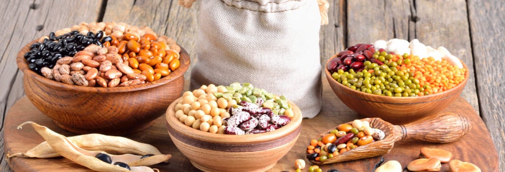 a variety of legumes and pulses organized in bags and bowls on a wooden tray, including black beans, cranberry beans, kidney beans, white beans, split peas, mung beans, yellow lentils, red lentils, green lentils