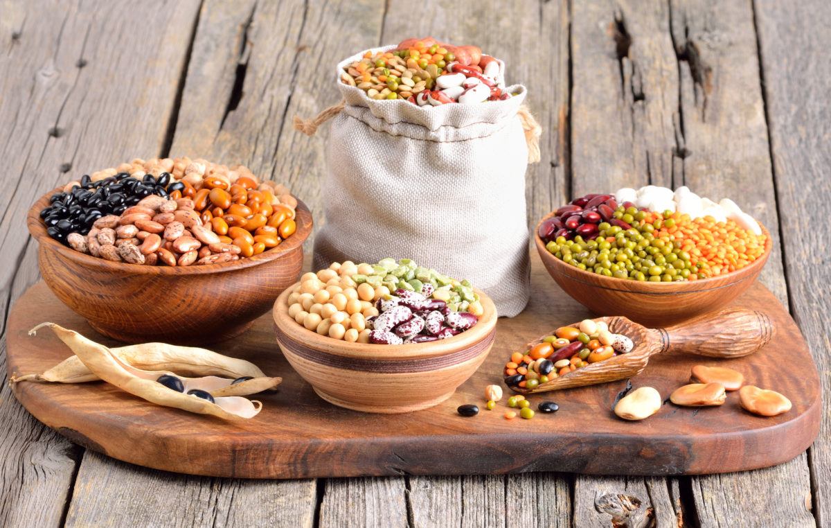 a variety of legumes and pulses organized in bags and bowls on a wooden tray, including black beans, cranberry beans, kidney beans, white beans, split peas, mung beans, yellow lentils, red lentils, green lentils