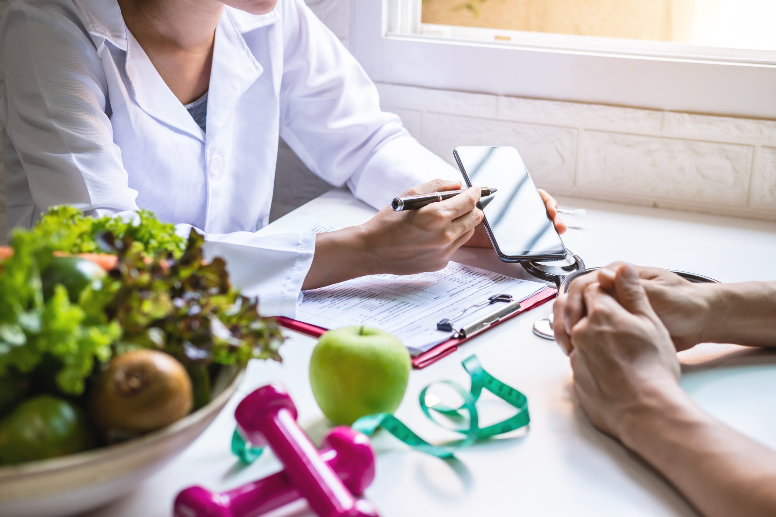 physician counseling a patient using a phone app on personalized nutrition recommendations. Sitting at a desk with various items including a bowl of vegetables, an apple, clipboard, and free weights