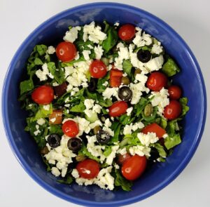 feta cheese sprinkled on top of a tomato, cucumber, olive, and lettuce salad in a blue bowl 