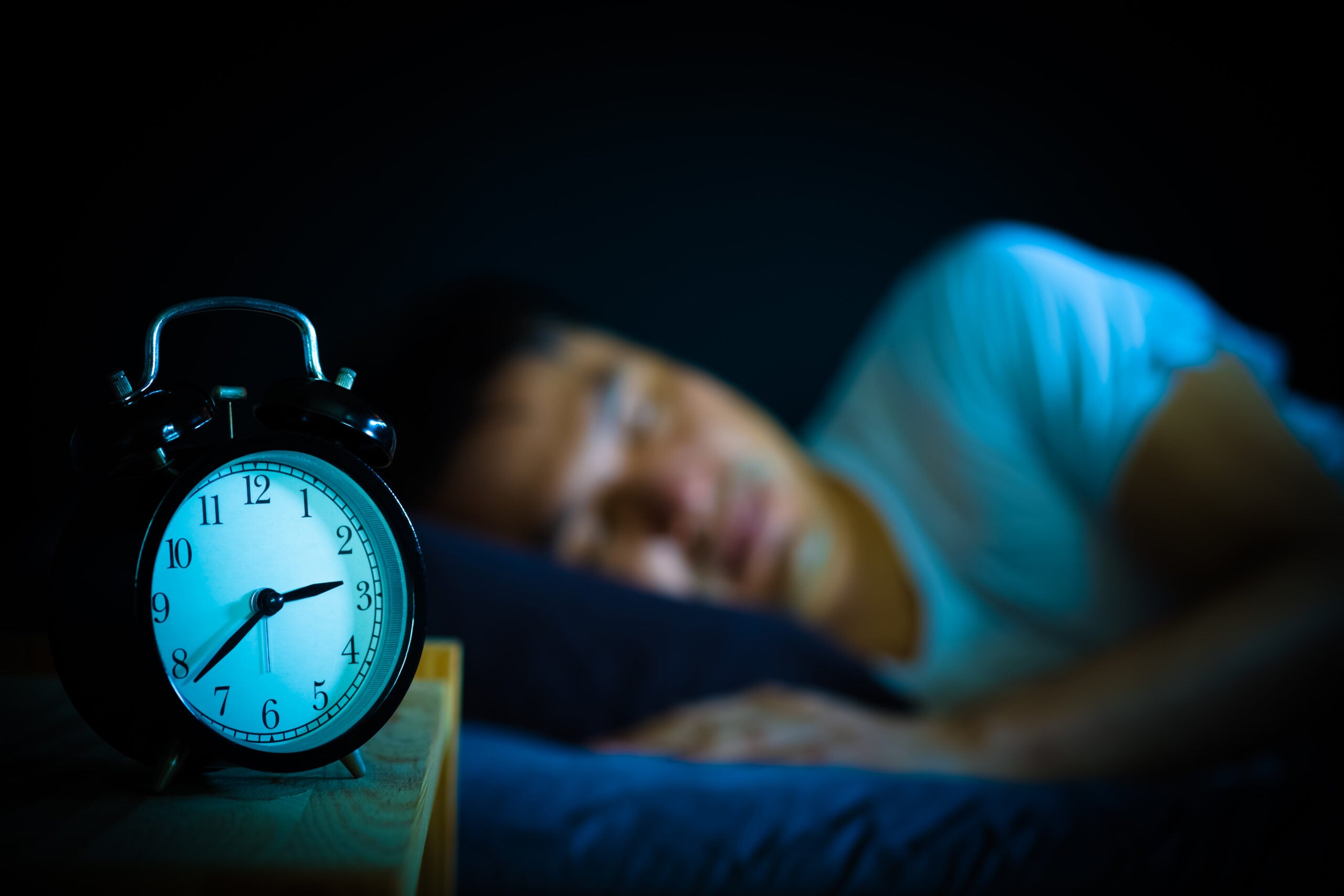 man sleeping in a bed at night with an alarm clock in the foreground showing the time as three am