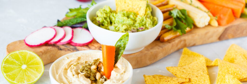 snacks arranged on a board including tortilla chips, hummus, guacamole, and a variety of sliced vegetables and fruits such as carrots, radishes, avocado, and figs
