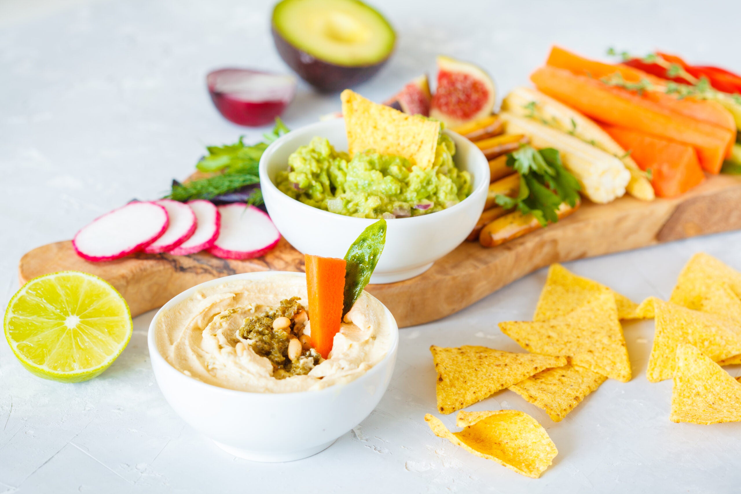 snacks arranged on a board including tortilla chips, hummus, guacamole, and a variety of sliced vegetables and fruits such as carrots, radishes, avocado, and figs
