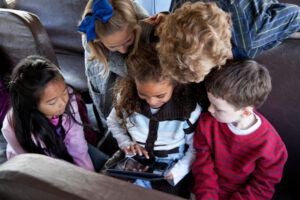 School children (ages 6 to 9) inside school bus huddled around girl playing with digital tablet.