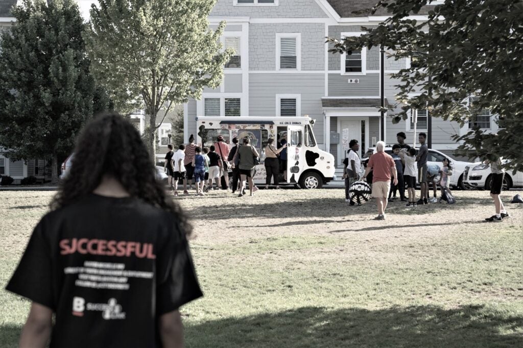 A girl with "successful" written on the back of her shirt walking towards an ice cream truck in a park surrounded by a crowd of people.