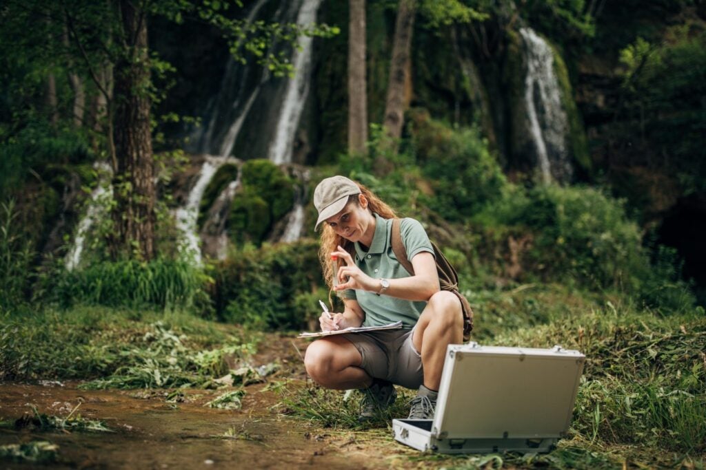 Woman sitting by river analyzing water sample.