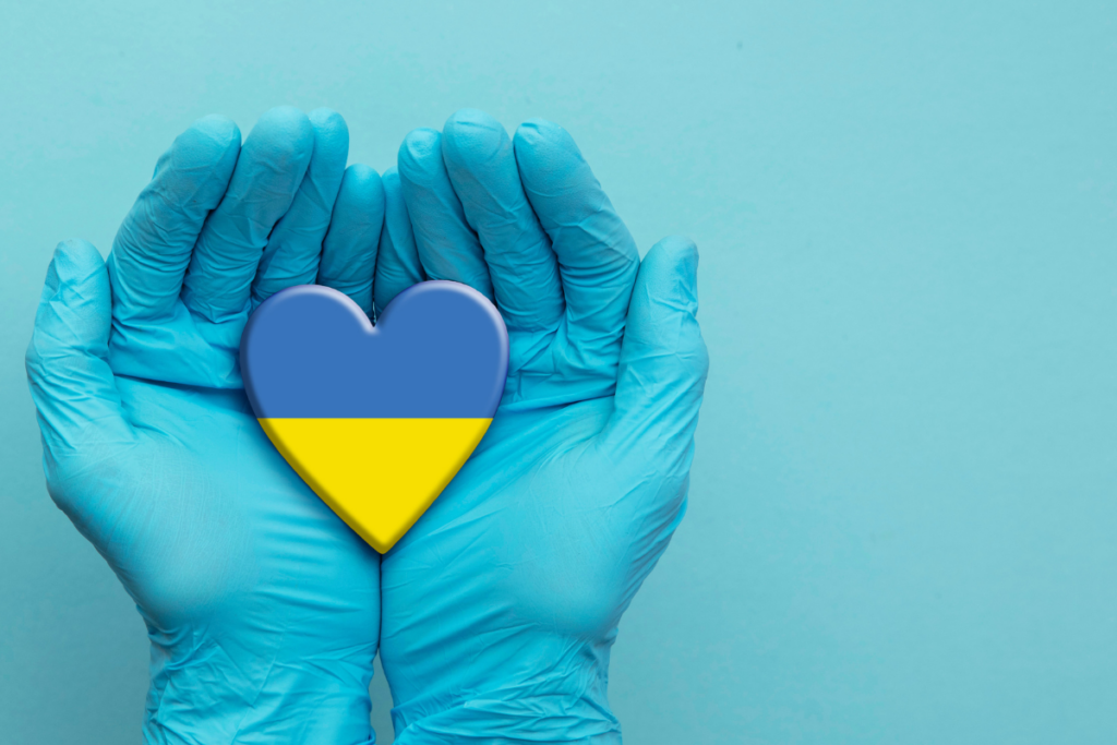 A pair of hands in blue surgical gloves cradles a hear in the colors of the Ukrainian flag against a light blue background.