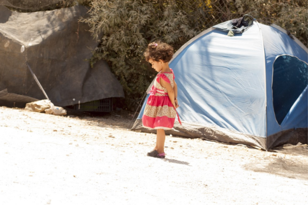 Young girl in dress standing in front of a blue tent at a refugee camp in Greece.