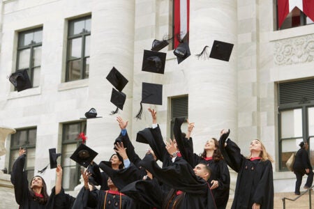 A group of Harvard Chan School Graduates stand in front of a large building wearing black graduation robes. The students are throwing their graduation caps into the air.