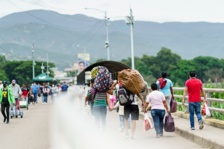 Photo of people carrying backpacks and bags walking across bridge. Mountains in the distance.