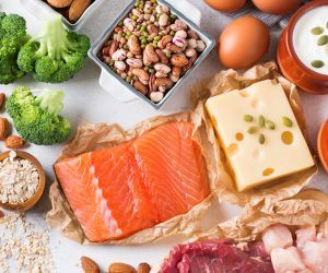 A variety of protein foods, including egg, salmon, beef, chicken, beans, lentils, almonds, quinoa, oats, broccoli, artichokes, yogurt, cheese, and tofu